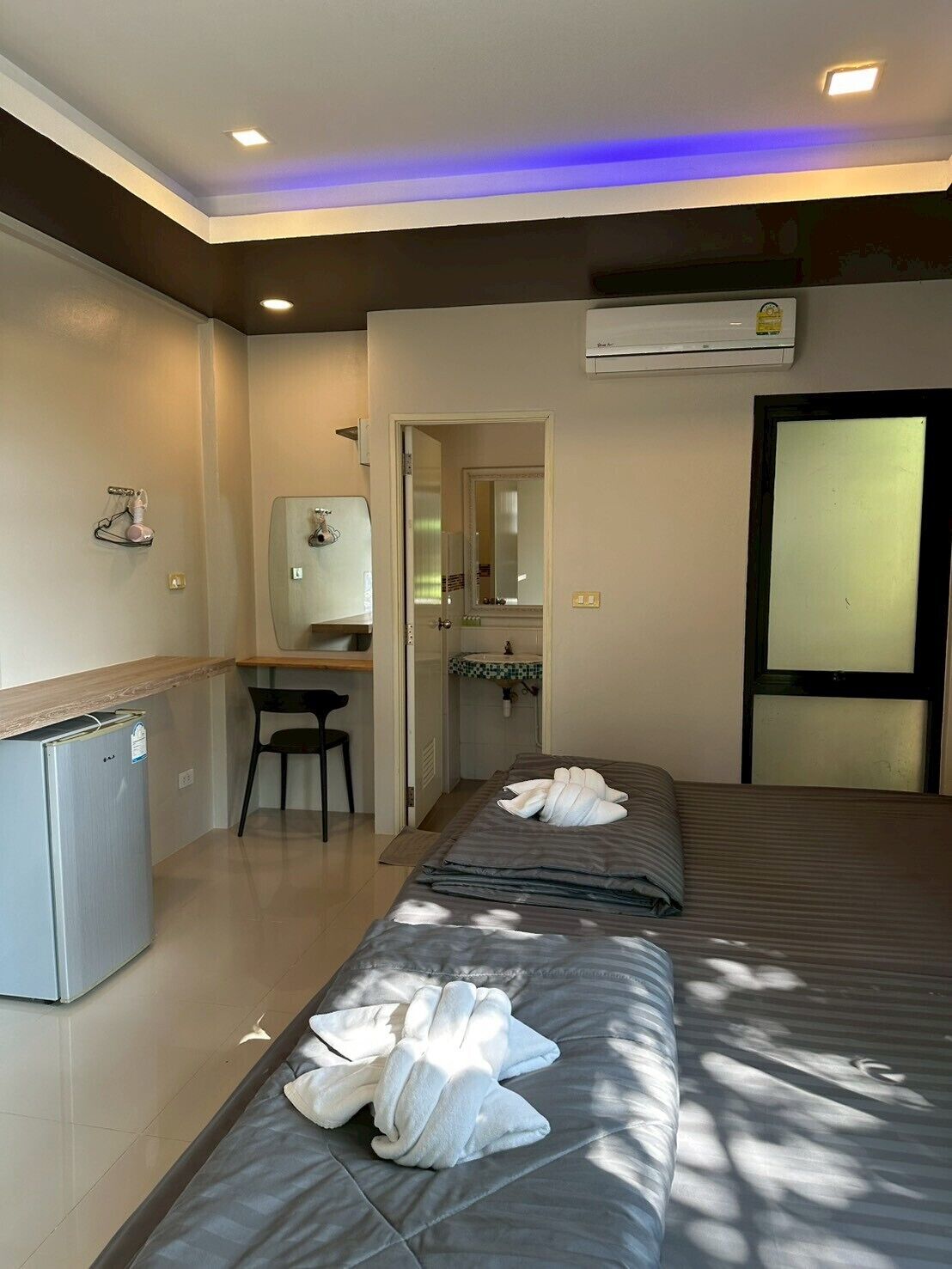 Habitación de hotel con dos camas, un escritorio, un refrigerador pequeño y un baño visible a través de una puerta abierta.