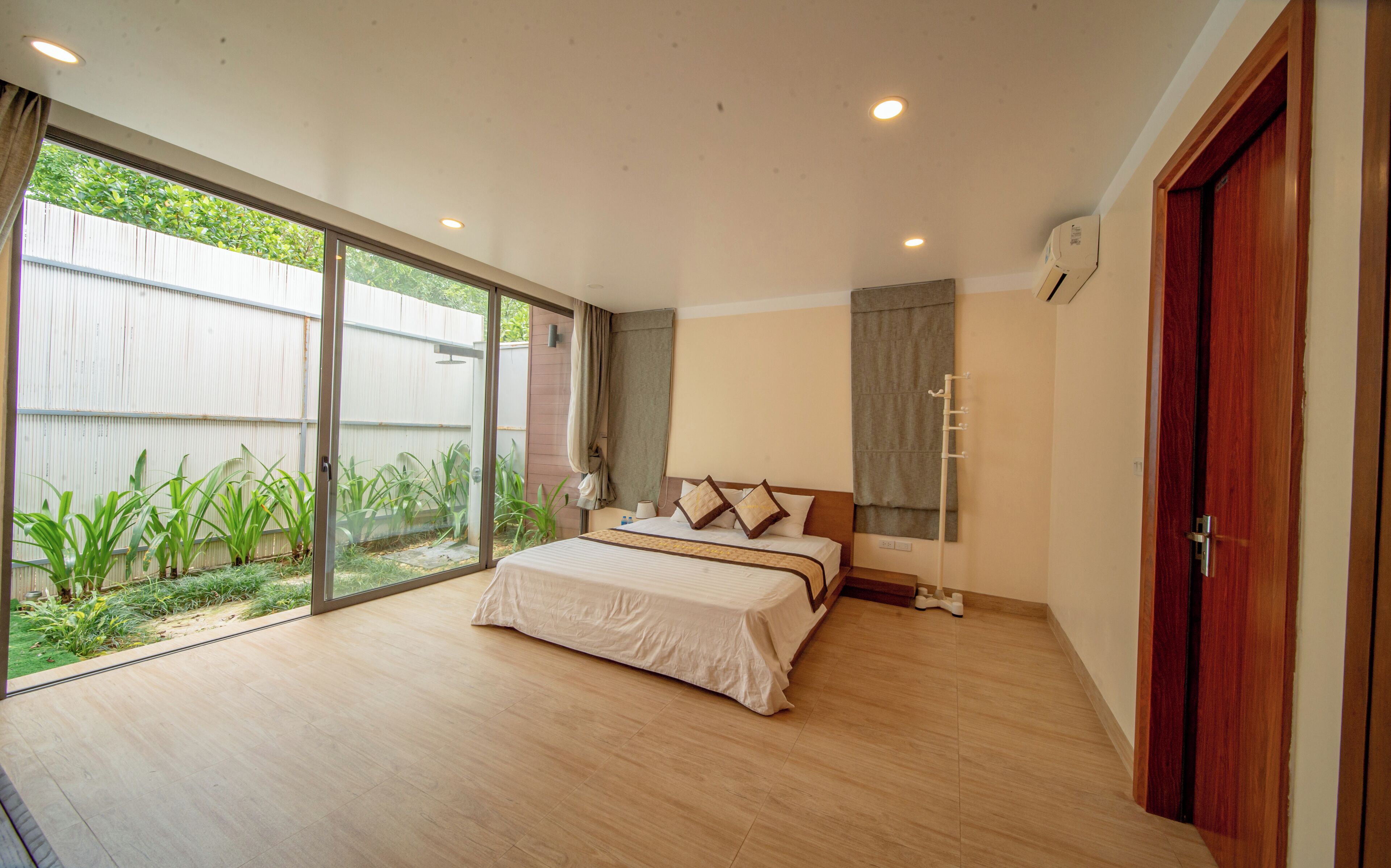 A bedroom with a large bed, sliding glass doors to a garden, a wooden floor, and a wooden door.