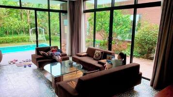 A spacious living room with a large glass table, two sofas, and a view of a swimming pool and greenery outside.