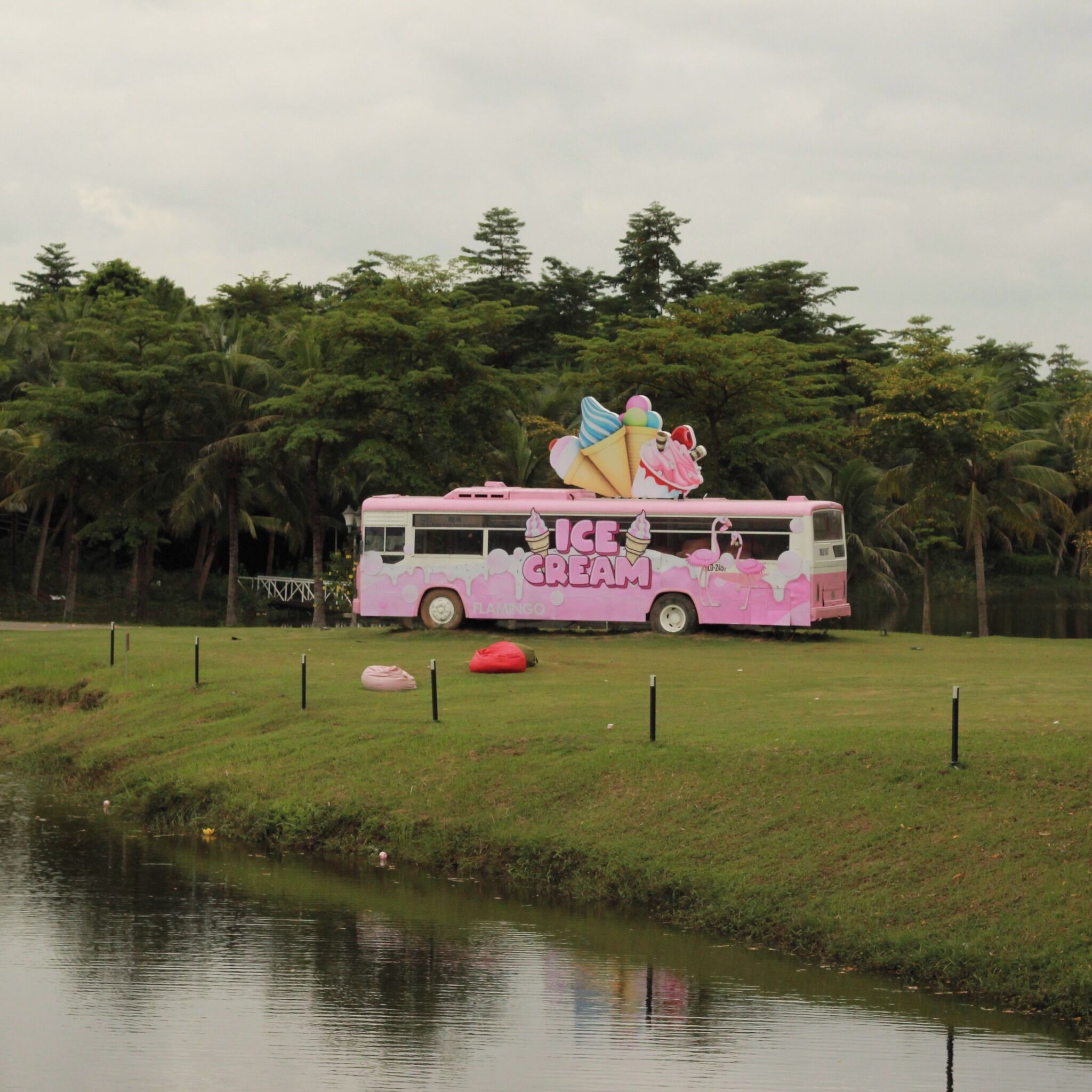Ein pinkfarbener Bus mit der Aufschrift 'ICE CREAM' und 'FLAMINGOS', geparkt auf einer Wiese in der Nähe von Wasser.