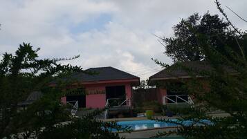 Zona de piscina con dos edificios, uno de fachada roja y otro de fachada rosa, rodeada de vegetación.