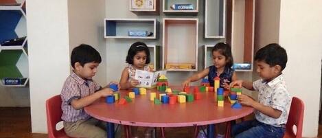 Four children playing with colorful blocks at a round table with red chairs.