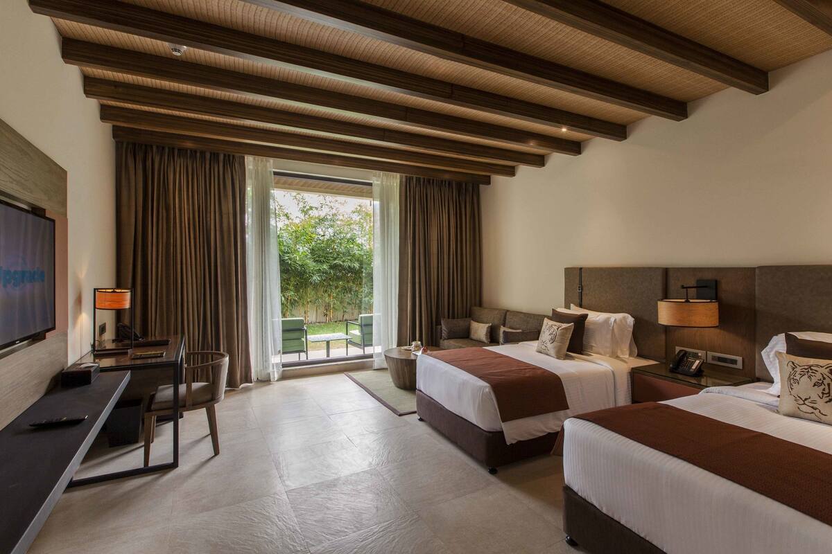 A hotel room with two beds, a desk, a TV, and a view of greenery outside.