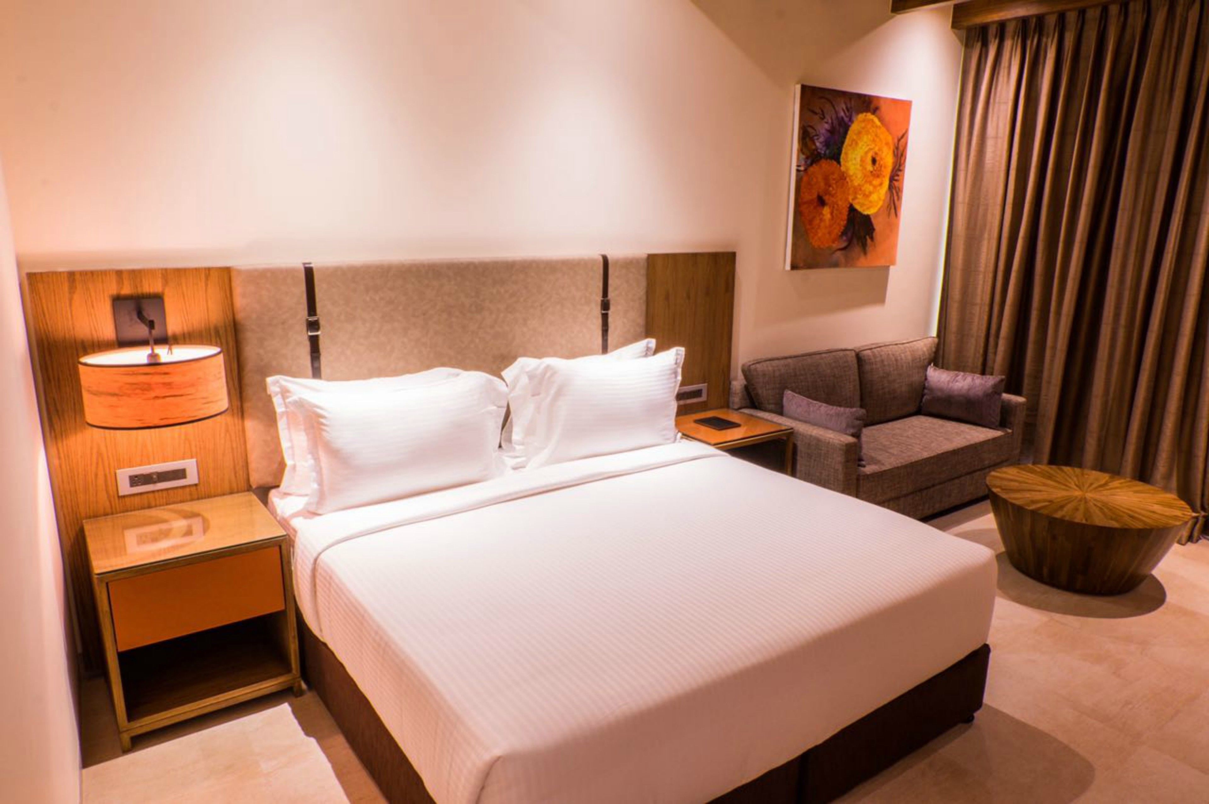 A hotel room with a large bed, a nightstand with a lamp, a small table, a sofa, and a decorative wall piece.