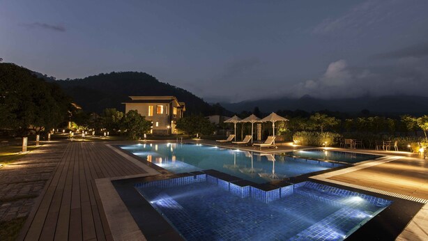 A nighttime pool area with a wooden deck, lounge chairs, and umbrellas, surrounded by mountains and a building.