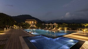 Un espace piscine aménagé pour la nuit, avec une terrasse en bois, des chaises longues et des parasols, le tout entouré de montagnes et d’un bâtiment.