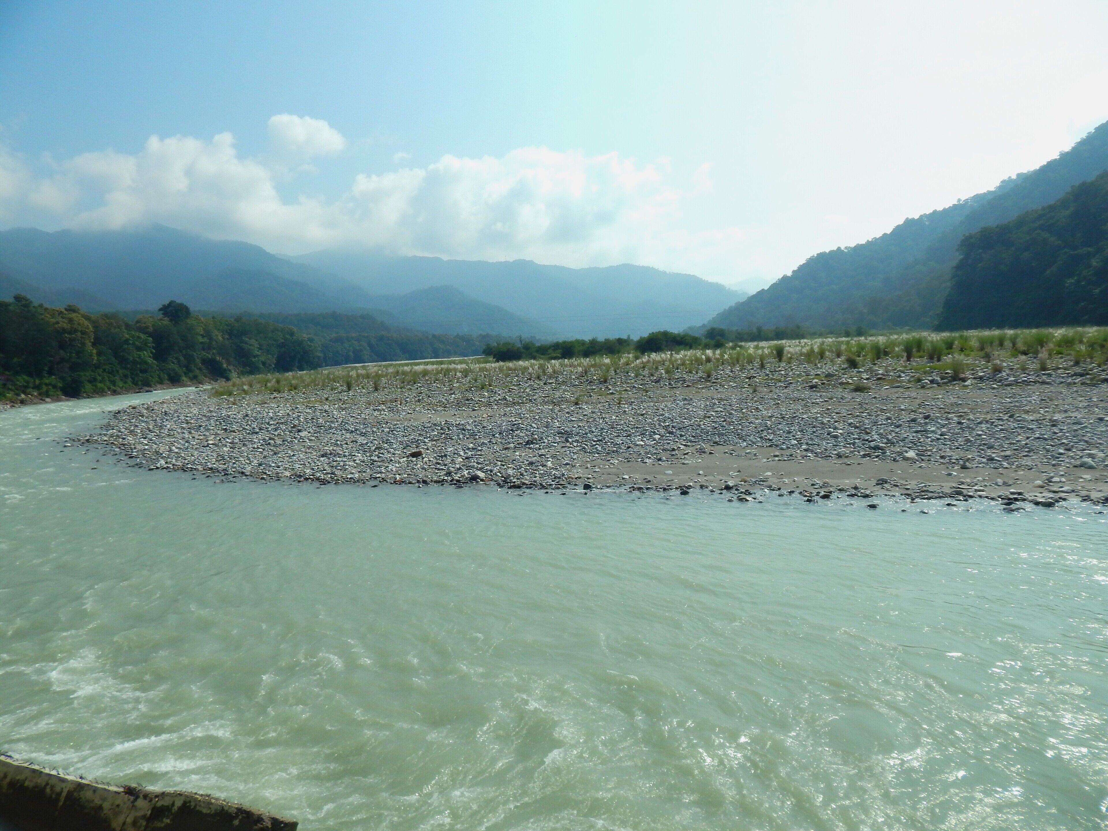 A river with a rocky riverbed, surrounded by lush greenery and distant mountains.