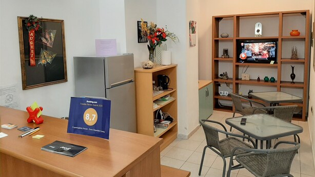 A reception area with a wooden desk, a refrigerator, a television, and a small seating area with chairs and a table. - Copenaghen House (Sassari)