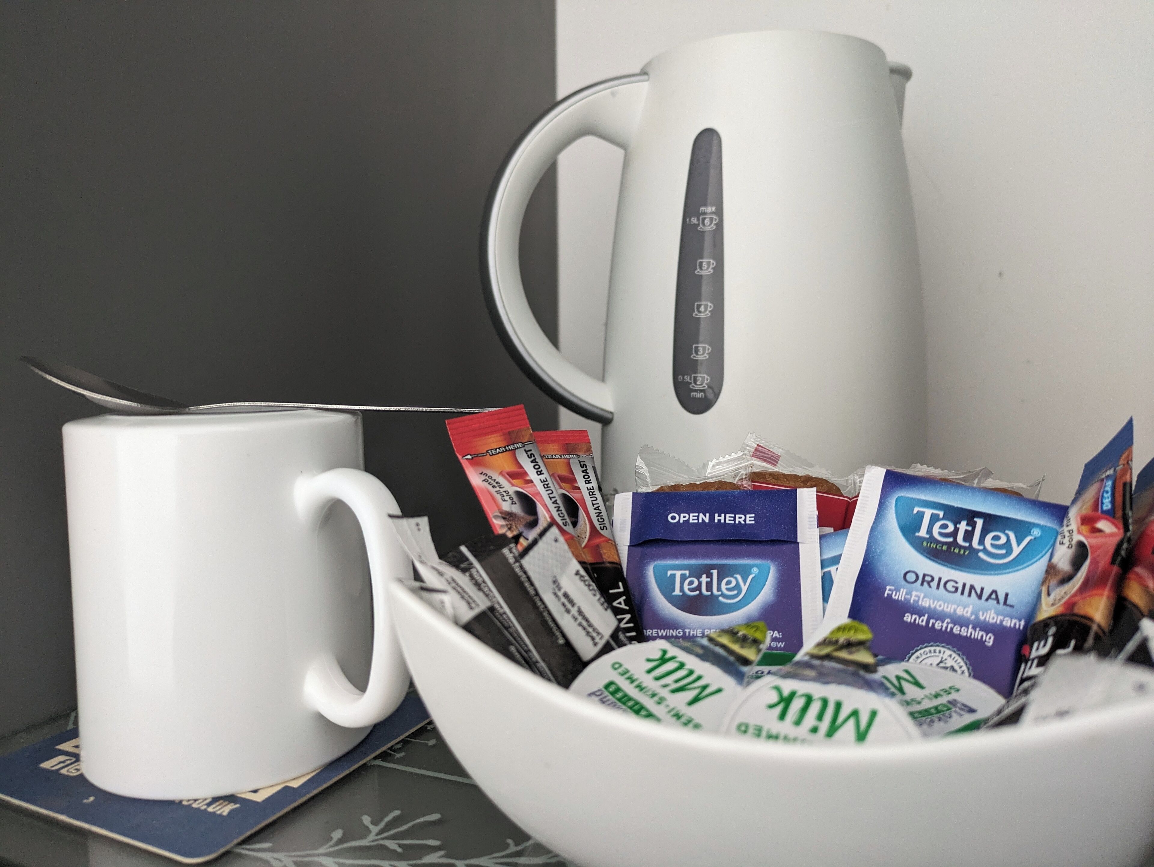 A white coffee maker, a white mug, and a bowl containing various coffee packets and a Tetley tea box.