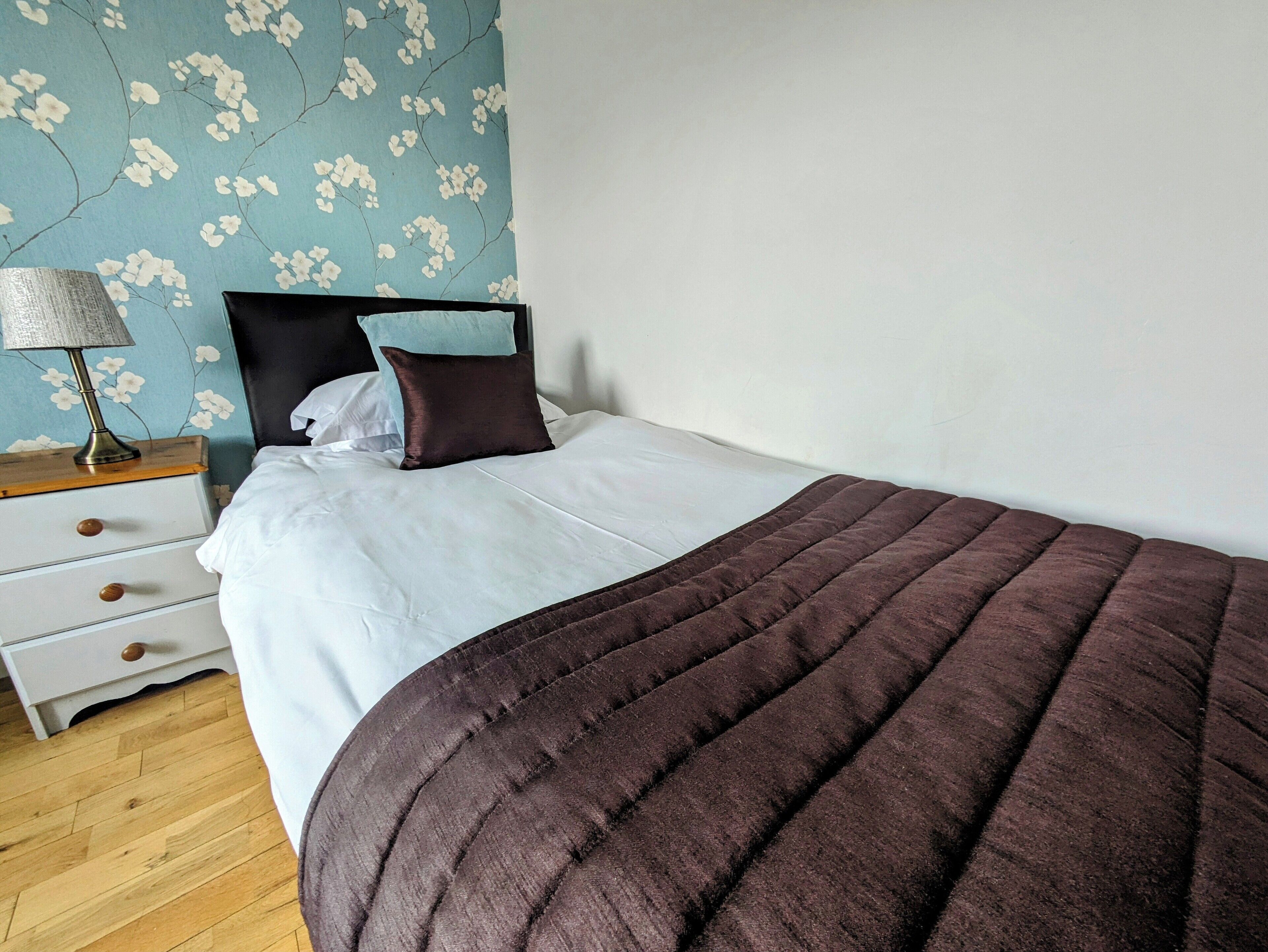 A bedroom with a bed, a nightstand, a lamp, and a floral wallpaper.