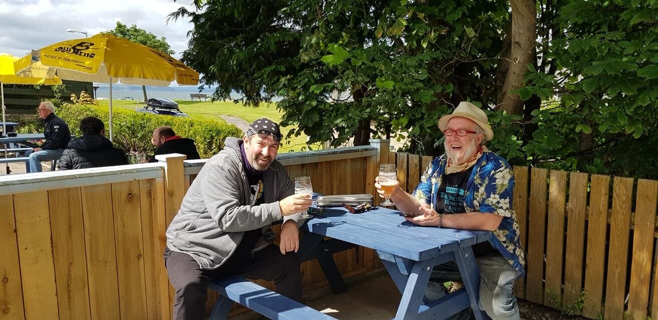 Dos personas sentadas en una mesa de madera al aire libre, con un paraguas amarillo al fondo.