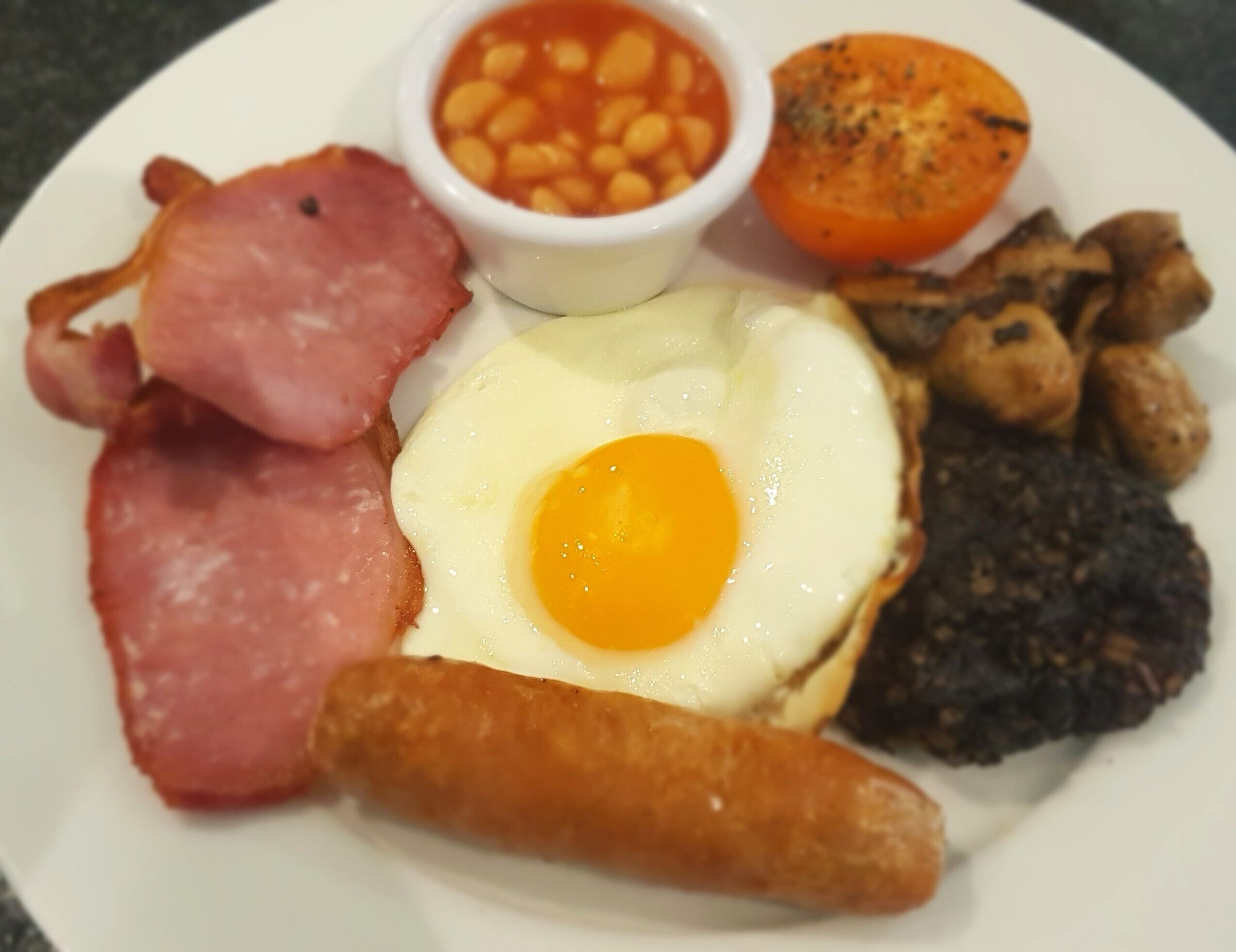 A plate with a fried egg, bacon, sausages, baked beans, a grilled tomato, and mushrooms.
