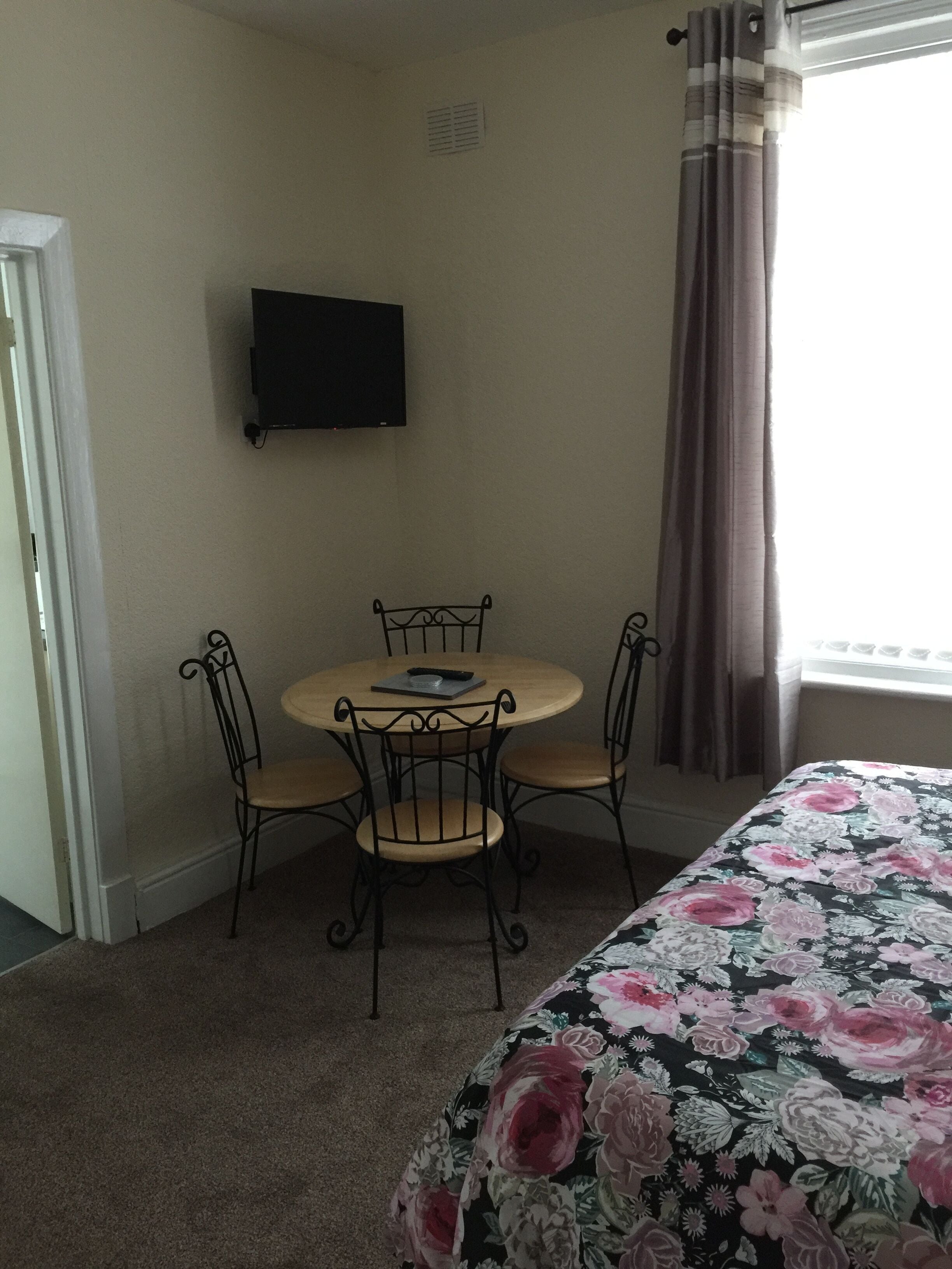 Una habitación pequeña con una cama, una mesa redonda con cuatro sillas, un televisor en la pared y una ventana con cortinas.