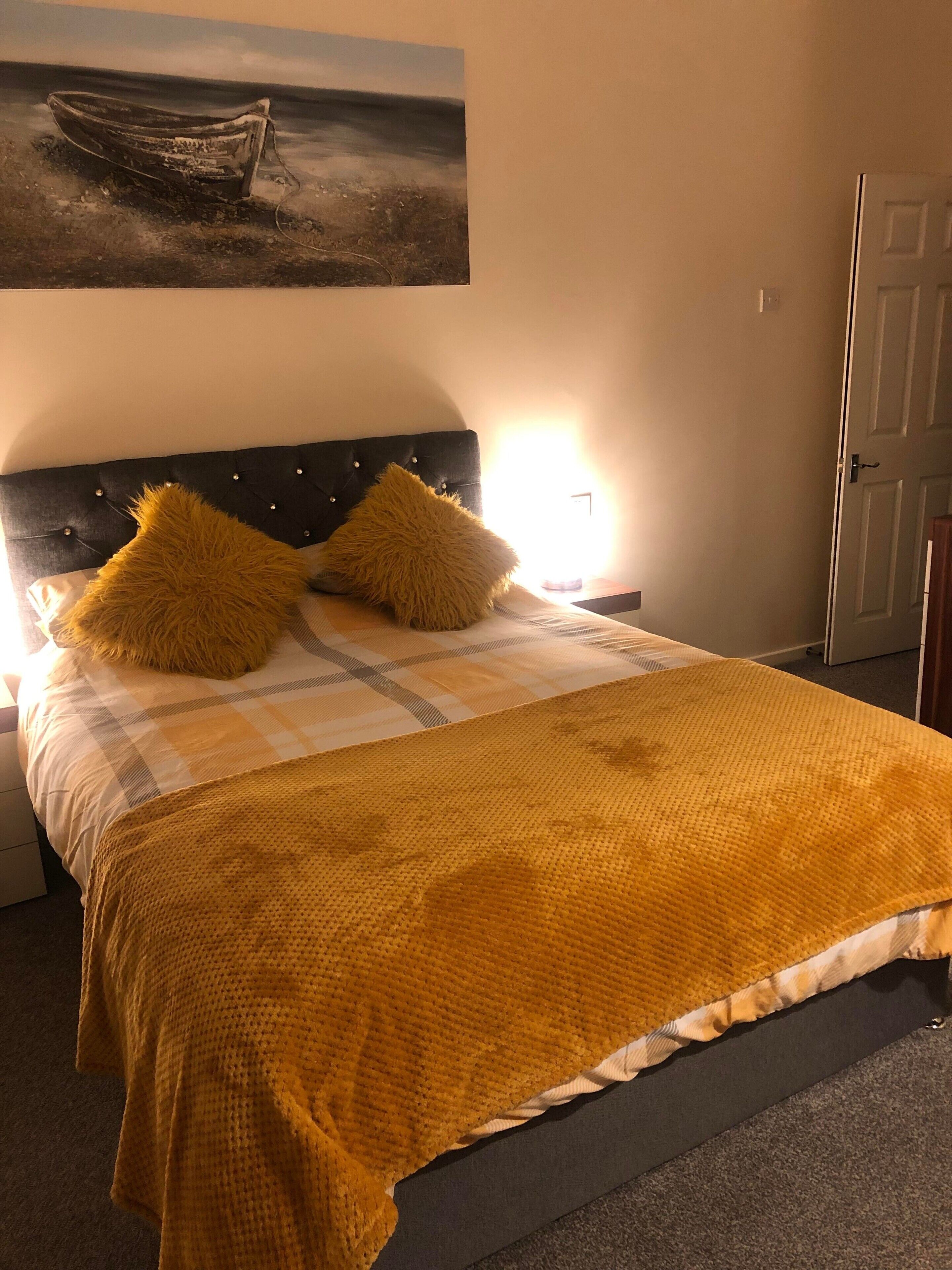 A bedroom with a bed, two pillows, a blanket, a headboard, a nightstand, a lamp, a door, and a painting on the wall.
