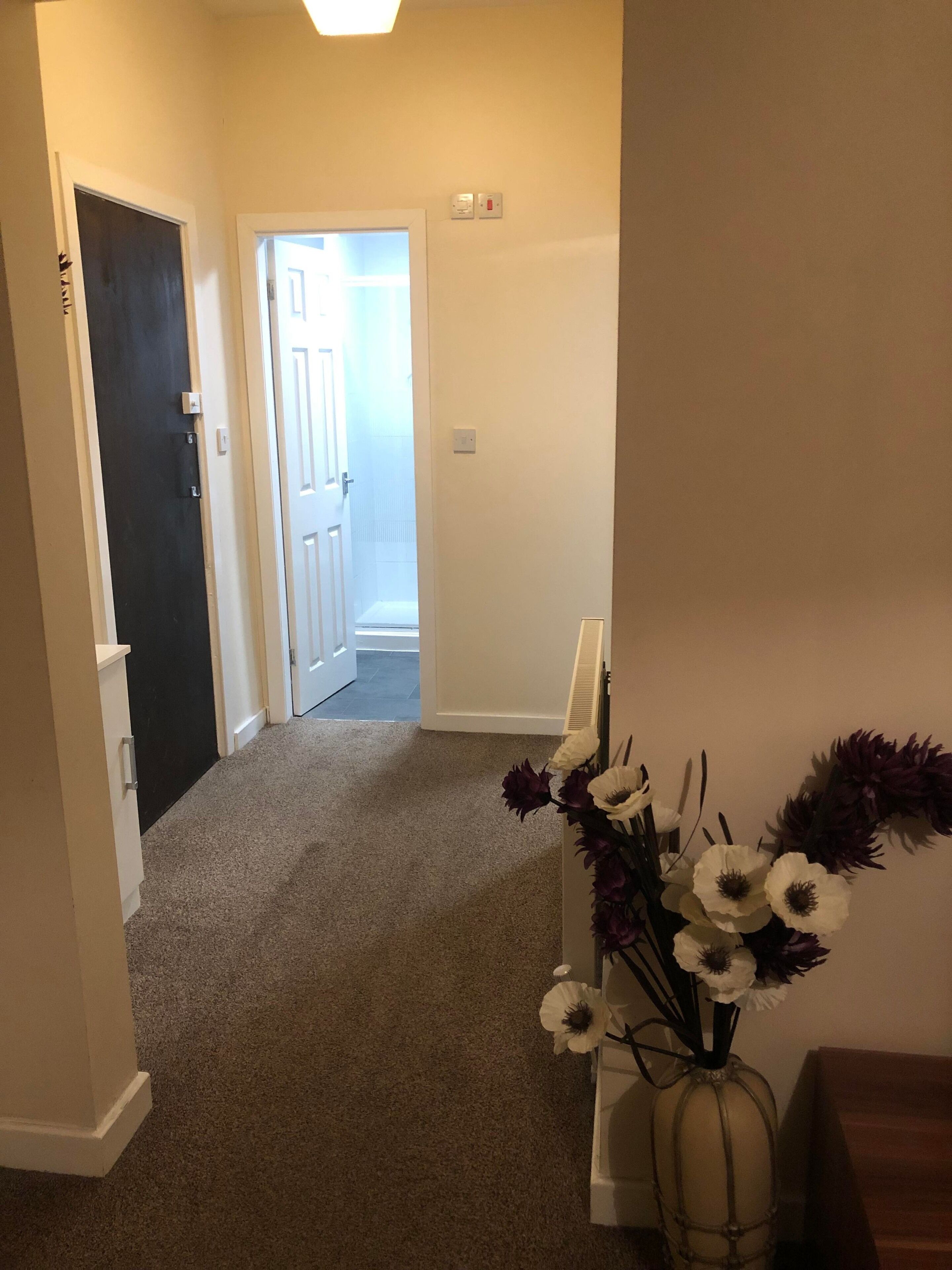A hallway with carpeted flooring, a dark door, and a white door leading to a stairwell. There is a vase with white and purple flowers on a white stand.