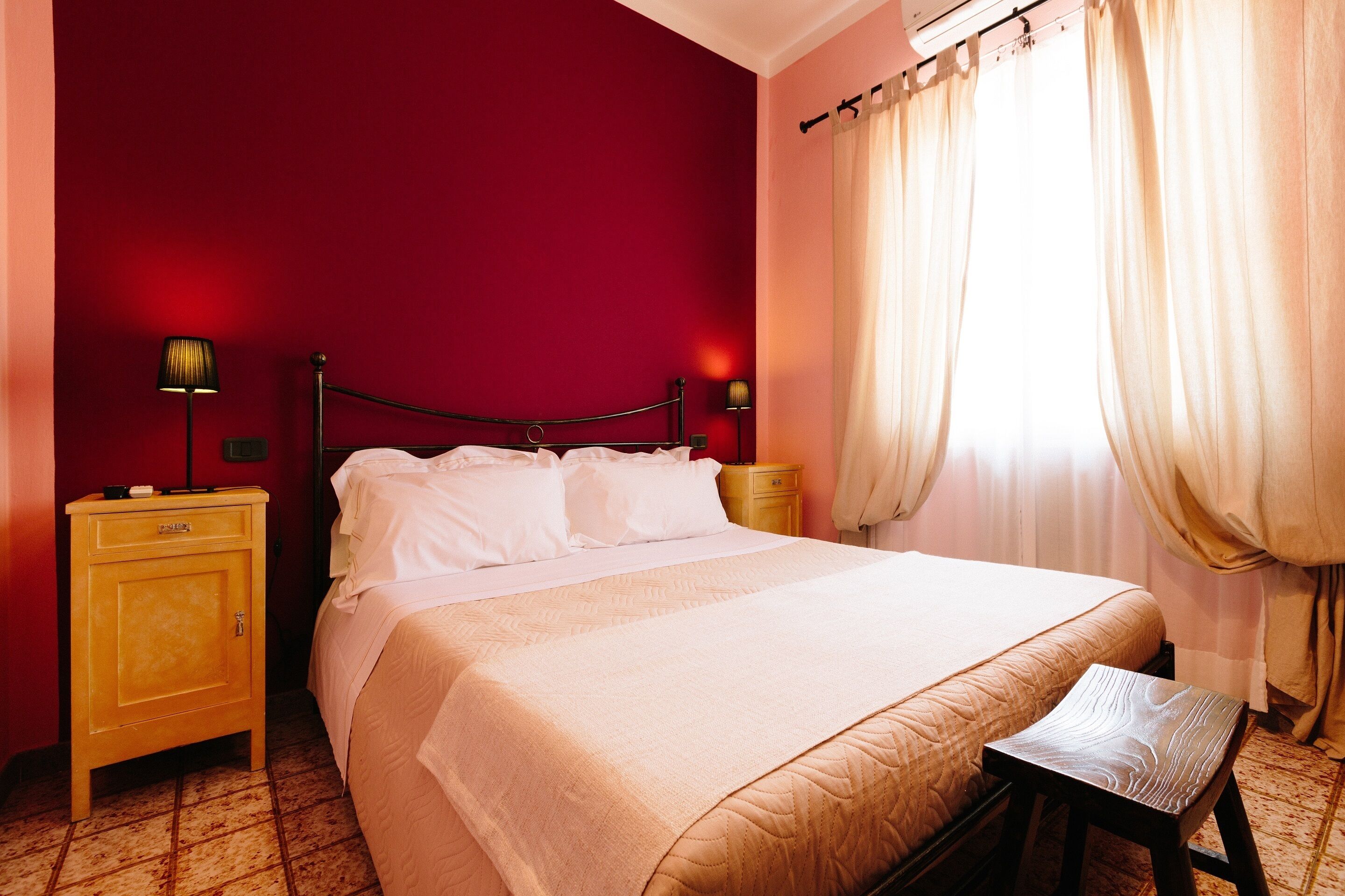 Una cama bien tendida con una colcha blanca y almohadas, una mesita de noche de madera con una lámpara, y un taburete de madera.