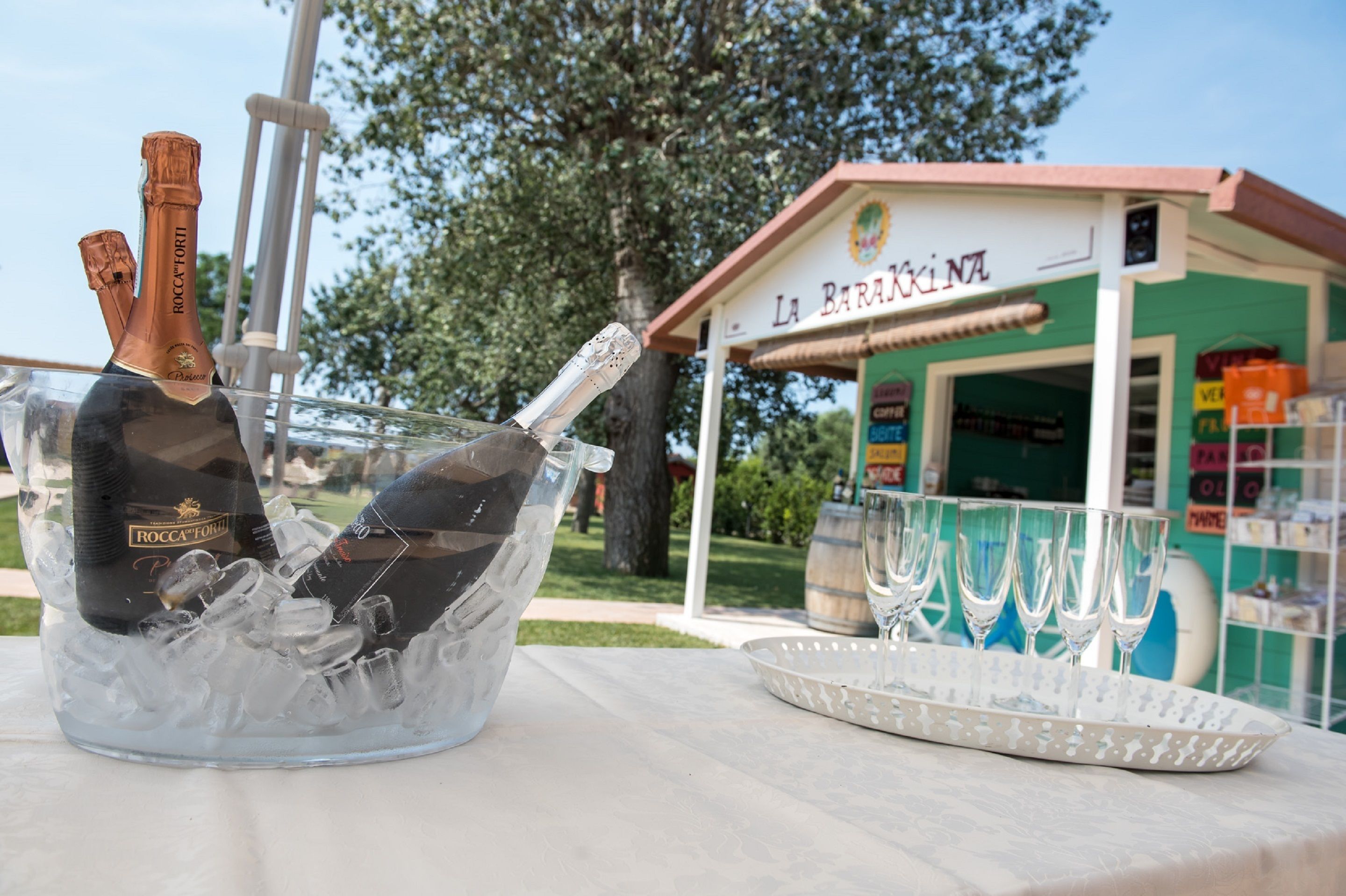 A bottle of Roccaforti champagne in a clear ice bucket, a tray with four wine glasses, and a storefront named La Barakina.