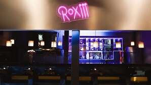 En bar med en neonreklamskylt som visar texten ”RoxIII” och en väl upplyst interiör med hyllor fulla av flaskor.