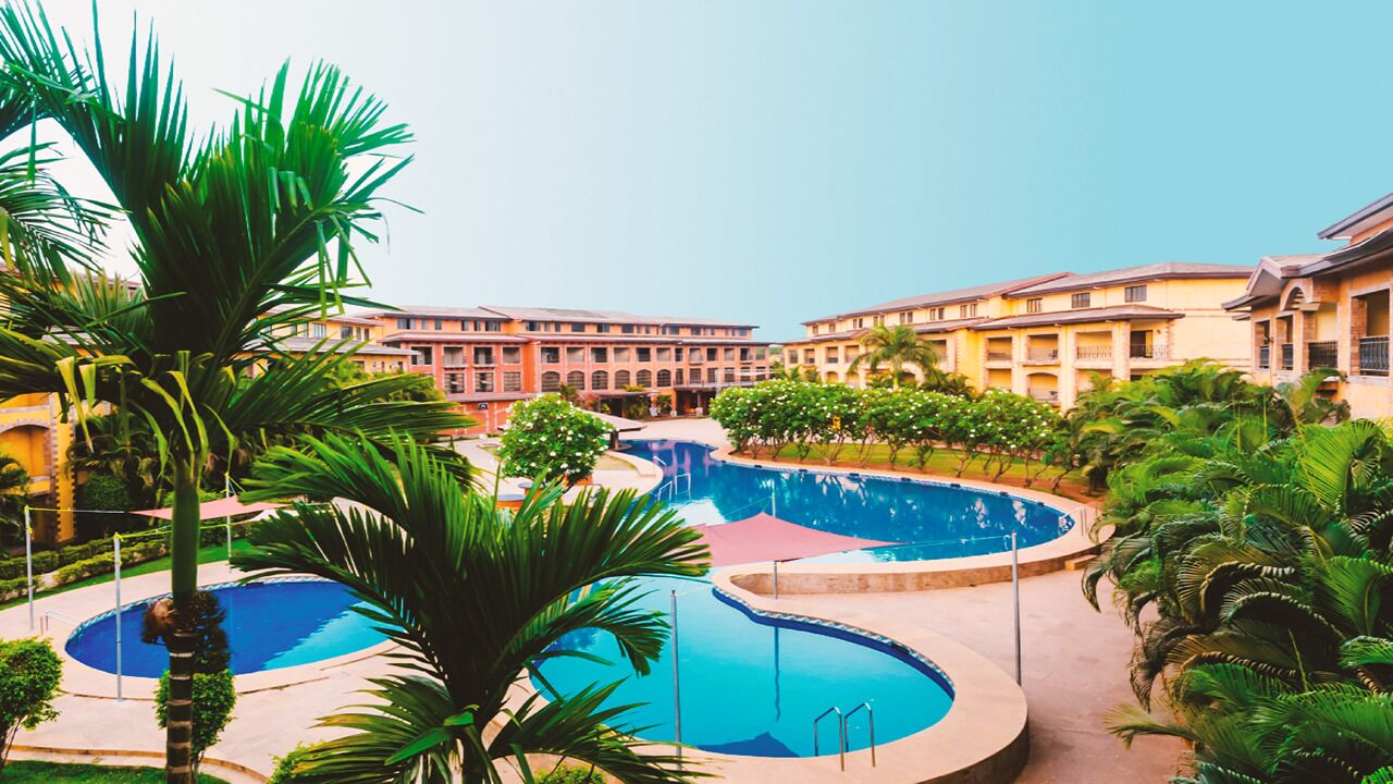 Et hotel med flere swimmingpools, palmer og en stor bygning med balkoner.