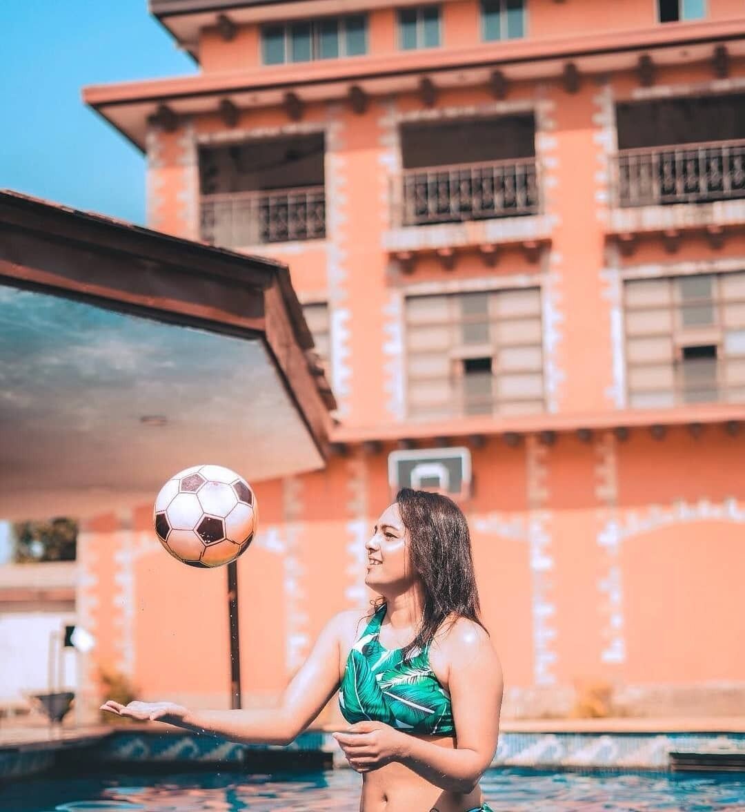 A person playing with a soccer ball by a pool with a building in the background.