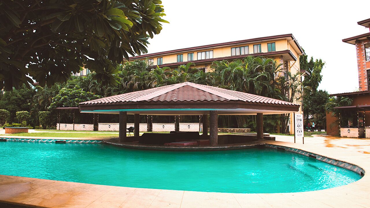 Piscina com um pavilhão coberto, cercada por vegetação exuberante e um edifício em vários andares ao fundo.