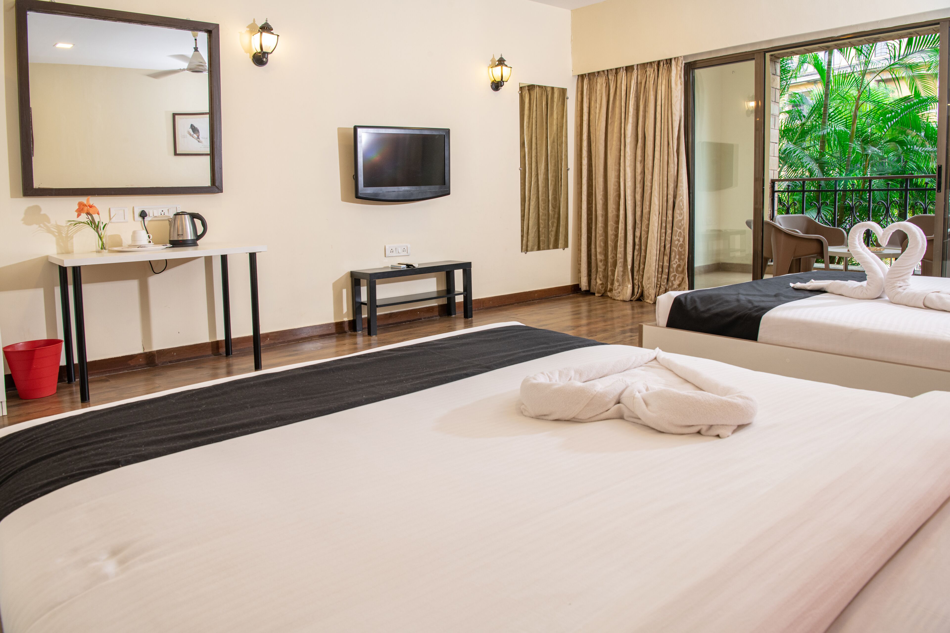 A hotel room with two beds, a TV, a desk with a kettle, a red bucket, and a balcony with a view of greenery.