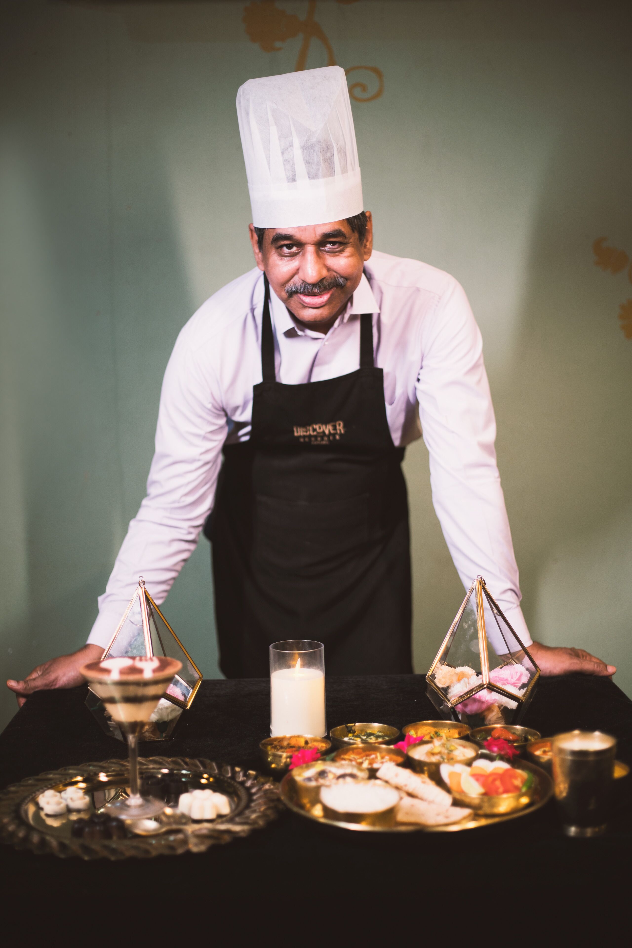 Une personne vêtue d’un uniforme de chef, debout derrière une table sur laquelle sont disposés divers plats et boissons.