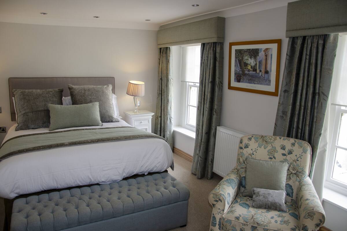 A bedroom with a large bed, a bedside table with a lamp, a chair, and a window with curtains.