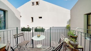 Een balkon met een witte muur, twee ramen, een tafel en enkele stoelen.