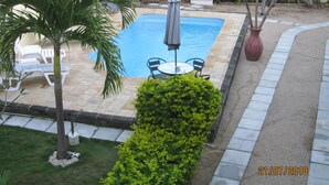 A swimming pool with lounge chairs and a table, surrounded by a garden with a hedge and palm trees. - Jolie Villa Santa (Pointe Aux Piments)