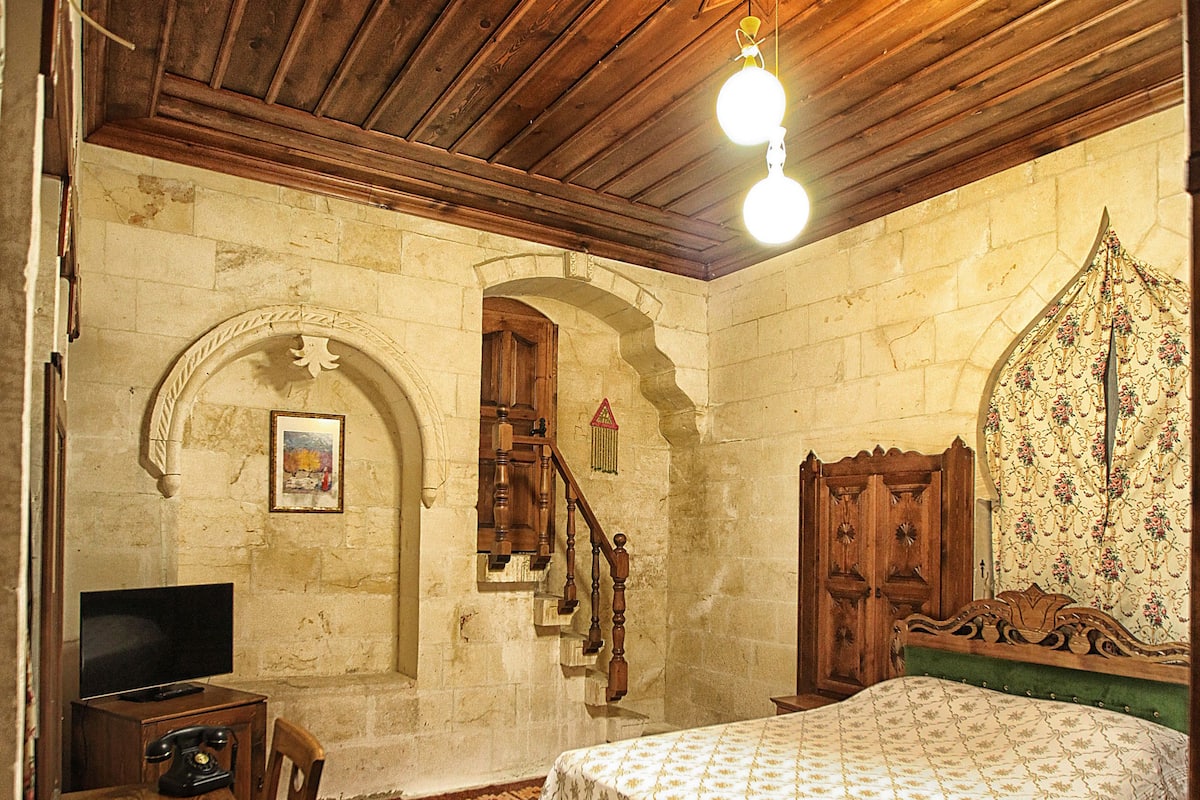 Una stanza con soffitto in legno, un letto con copripiumino decorato, una sedia di legno, un tavolino con un televisore e un arco decorativo sulla parete.