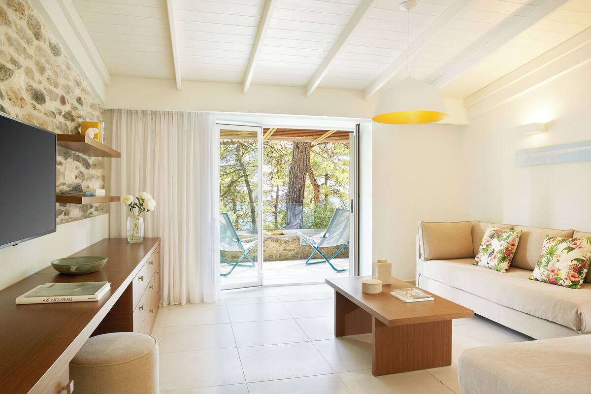 A modern living room with a sofa, coffee table, and a TV on a wooden cabinet. There is a sliding glass door leading to an outdoor area with chairs and a tree.
