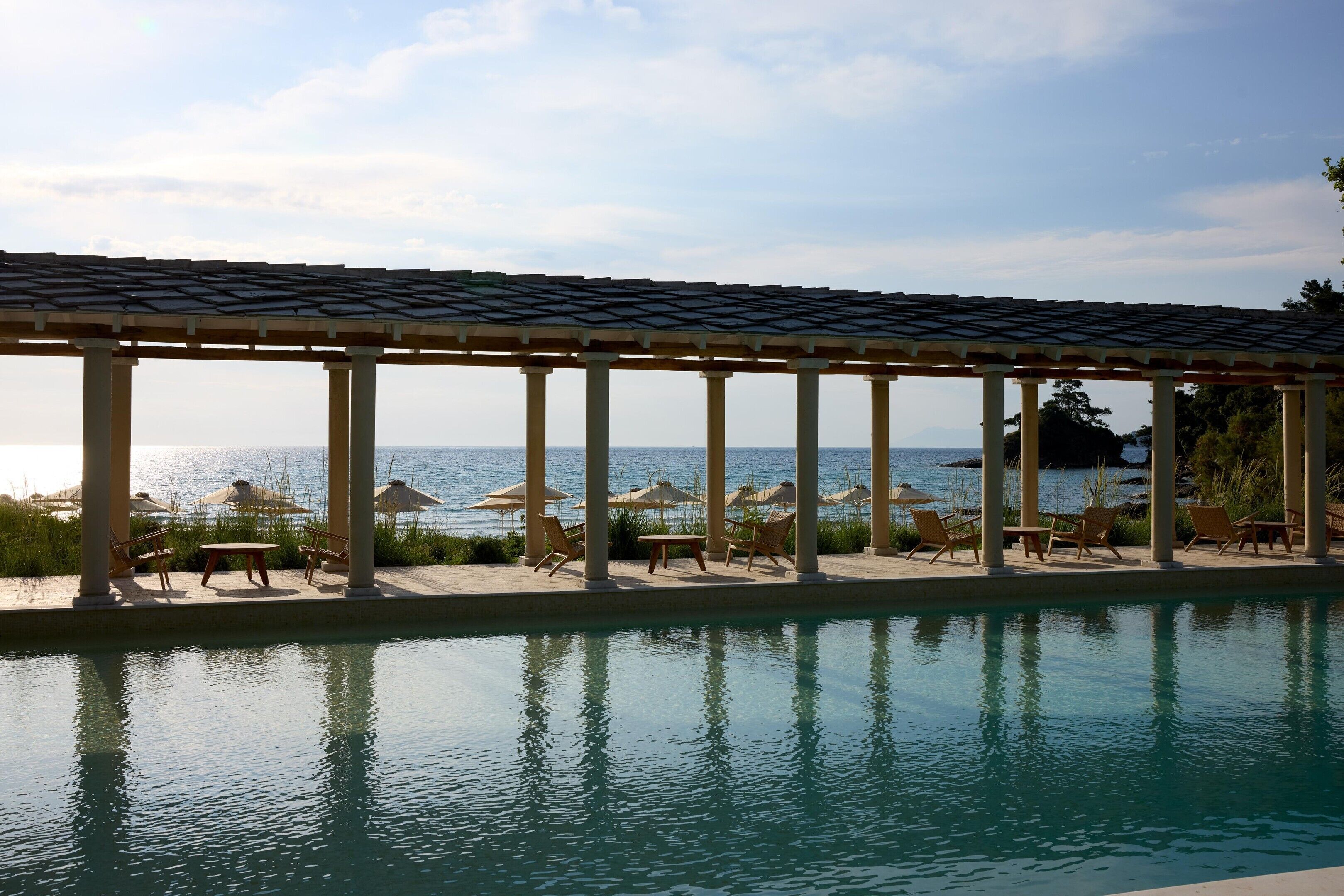 A pool area with a pergola, lounge chairs, and umbrellas overlooking a body of water.