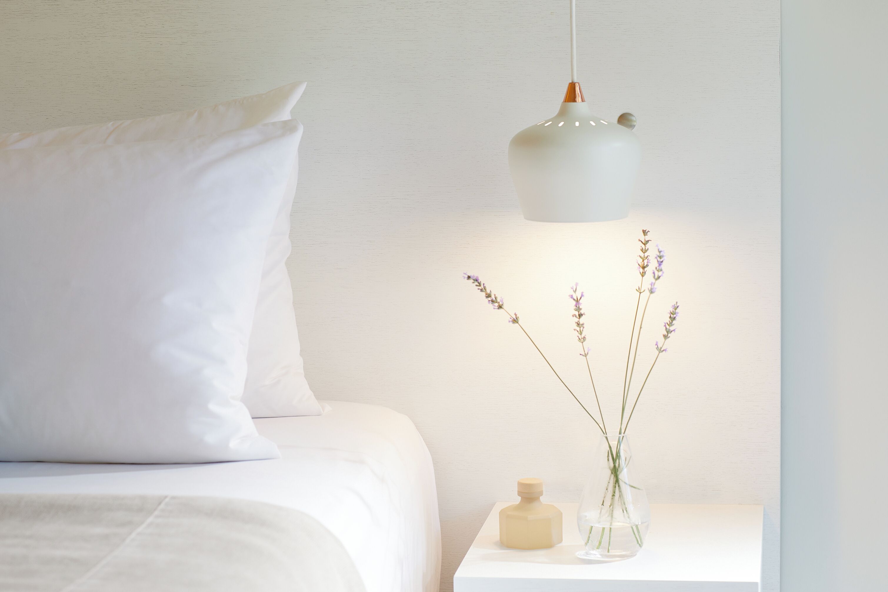 A neatly made bed with white bedding and pillows, a white bedside table with a vase of lavender flowers, and a modern pendant light.