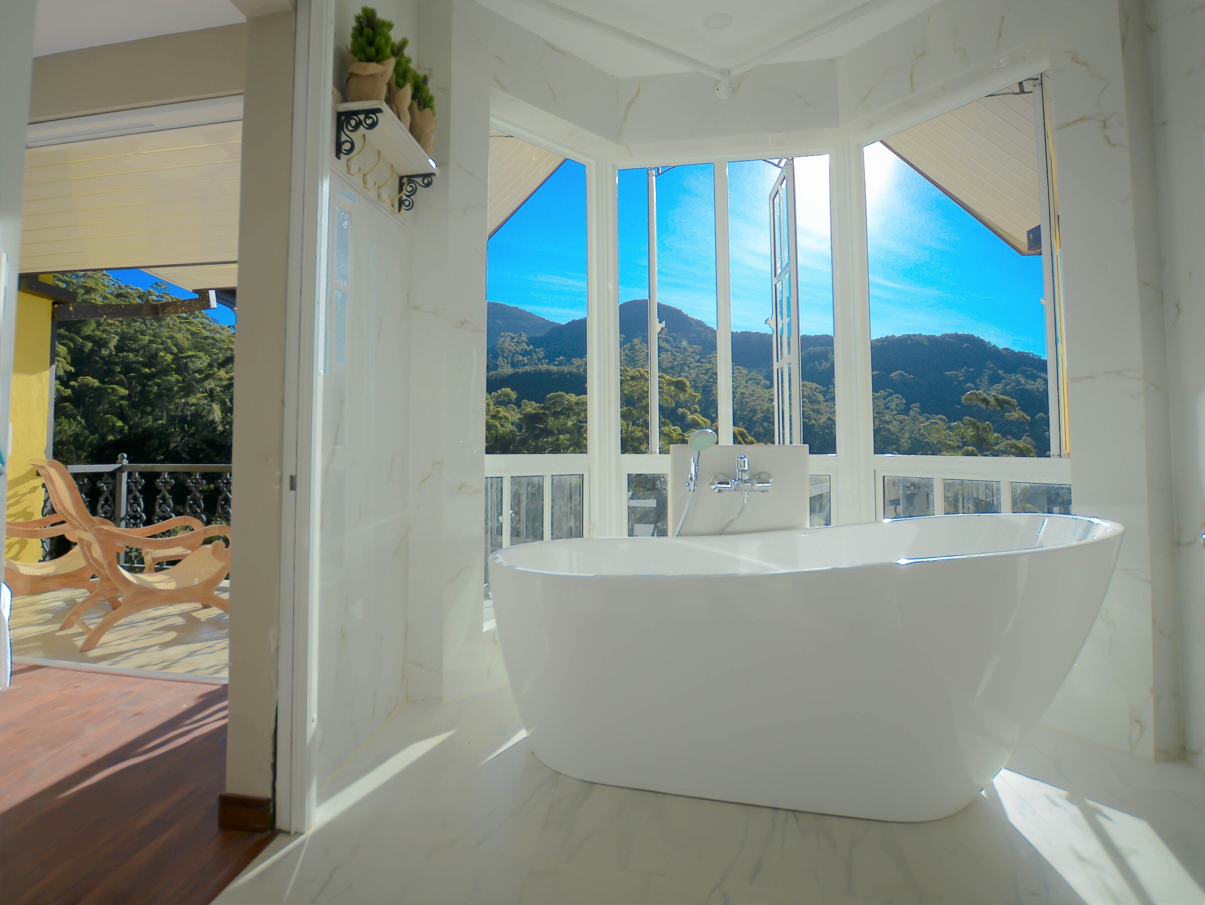 Un bagno moderno con una vasca da bagno freestanding, ampie finestre che offrono una vista panoramica su montagne e vegetazione, e un balcone con sedie in vimini.