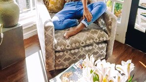 A woman sitting on a patterned armchair in a room with a large window, a lamp, and a vase of flowers. - Luxe Wilderness (Nuwara Eliya)