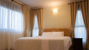 A hotel room with a large bed, beige curtains, and a wall-mounted light fixture. - Mayflower Serviced Apartments (Kampala)