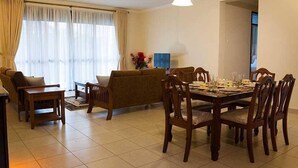 A dining area with a wooden table set for four, surrounded by chairs. There is a sofa and a small table with a vase of flowers. - Mayflower Serviced Apartments (Kampala)
