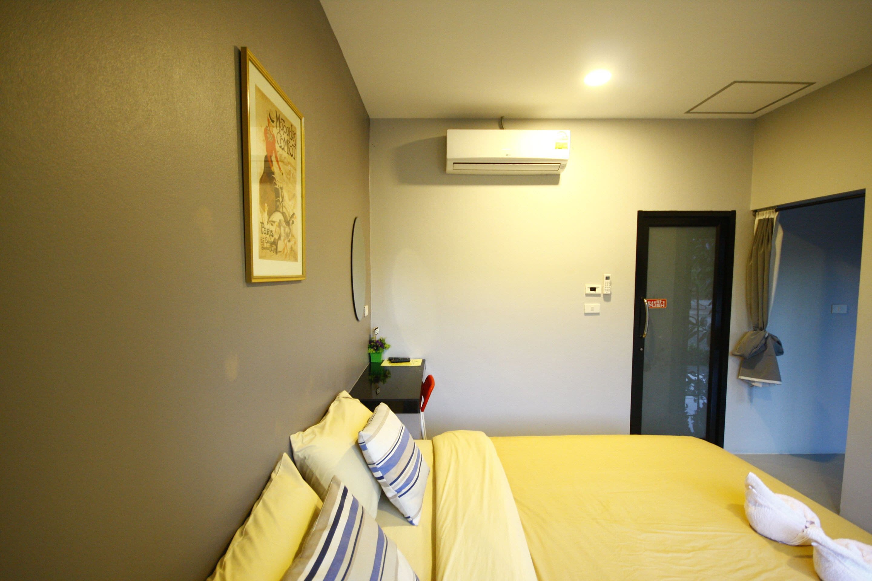 Quarto de hotel com cama, almofadas, ar condicionado, escrivaninha e uma cadeira, e porta.