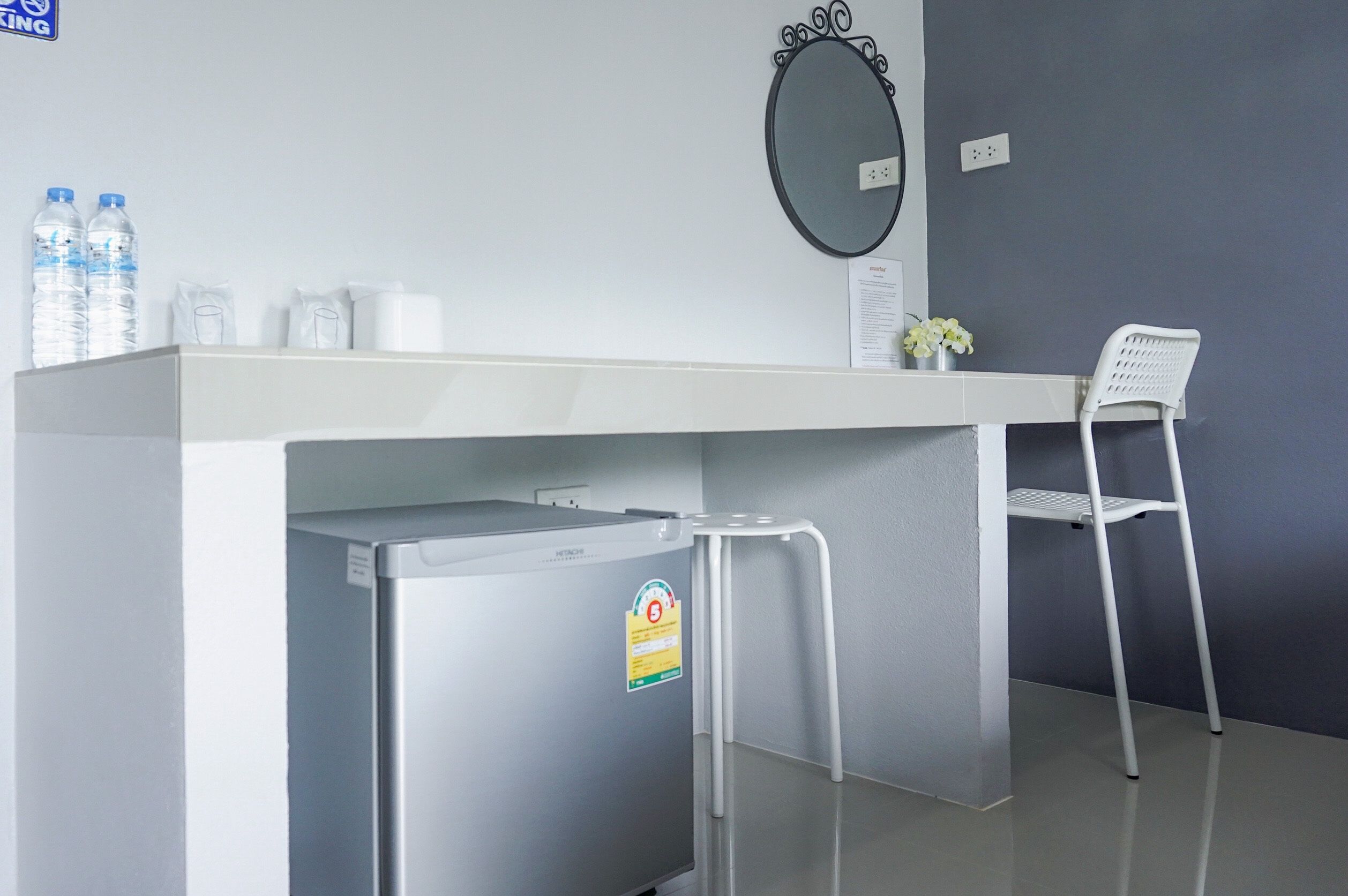 A compact hotel room with a white desk, a small refrigerator, a white chair, a mirror, and a water dispenser.