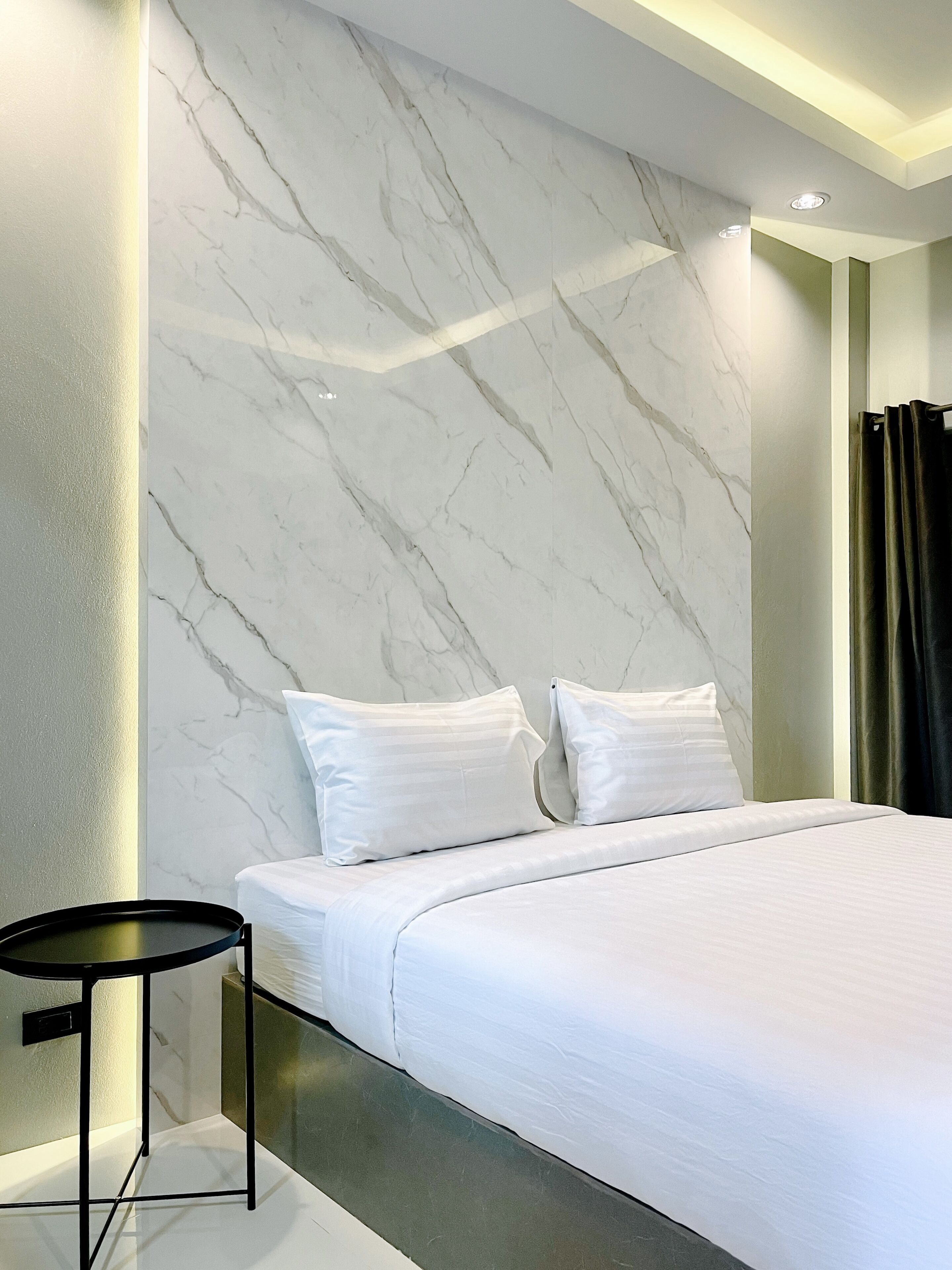Un dormitorio moderno con una cama grande, ropa de cama blanca, una pared con diseño de mármol y una pequeña mesa redonda.