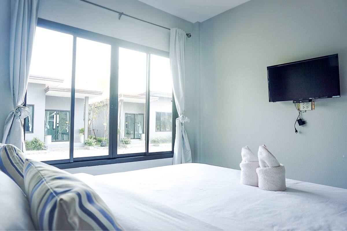 A hotel room with a large window, a bed with white linens, a flat-screen TV mounted on the wall, and a pair of towels on the bed.