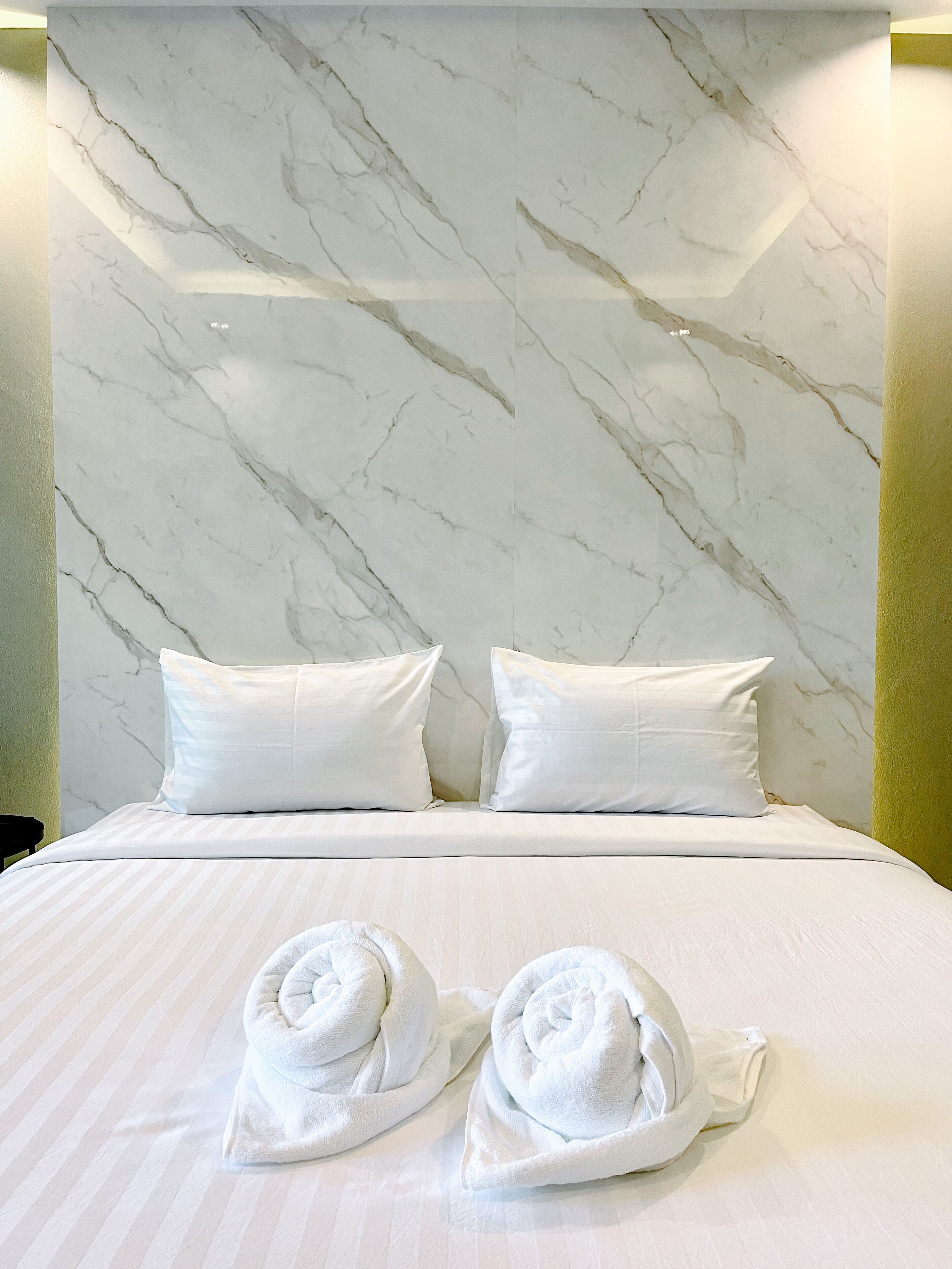 A hotel room with a marble headboard, two white pillows, and two rolled towels on the bed.