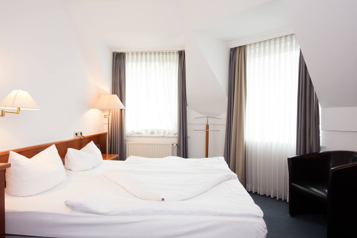 A hotel room with a large bed, two bedside tables with lamps, a chair, and a window with curtains.