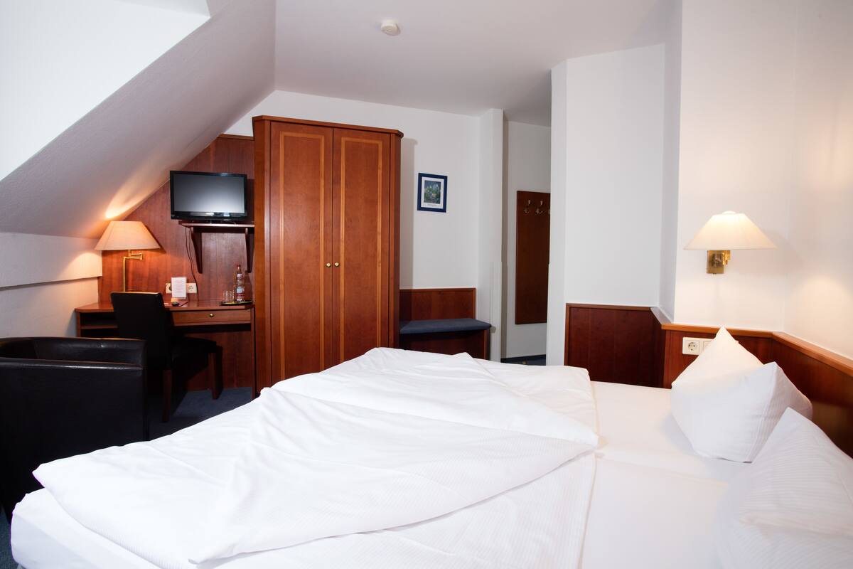 A hotel room with a bed, desk, chair, television, wardrobe, and a wall-mounted lamp.