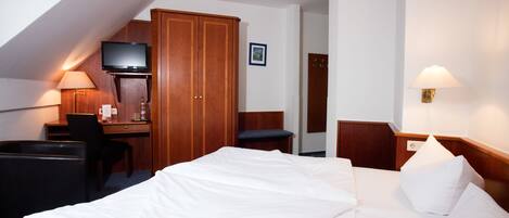 A hotel room with a bed, desk, chair, television, wardrobe, and a wall-mounted lamp.
