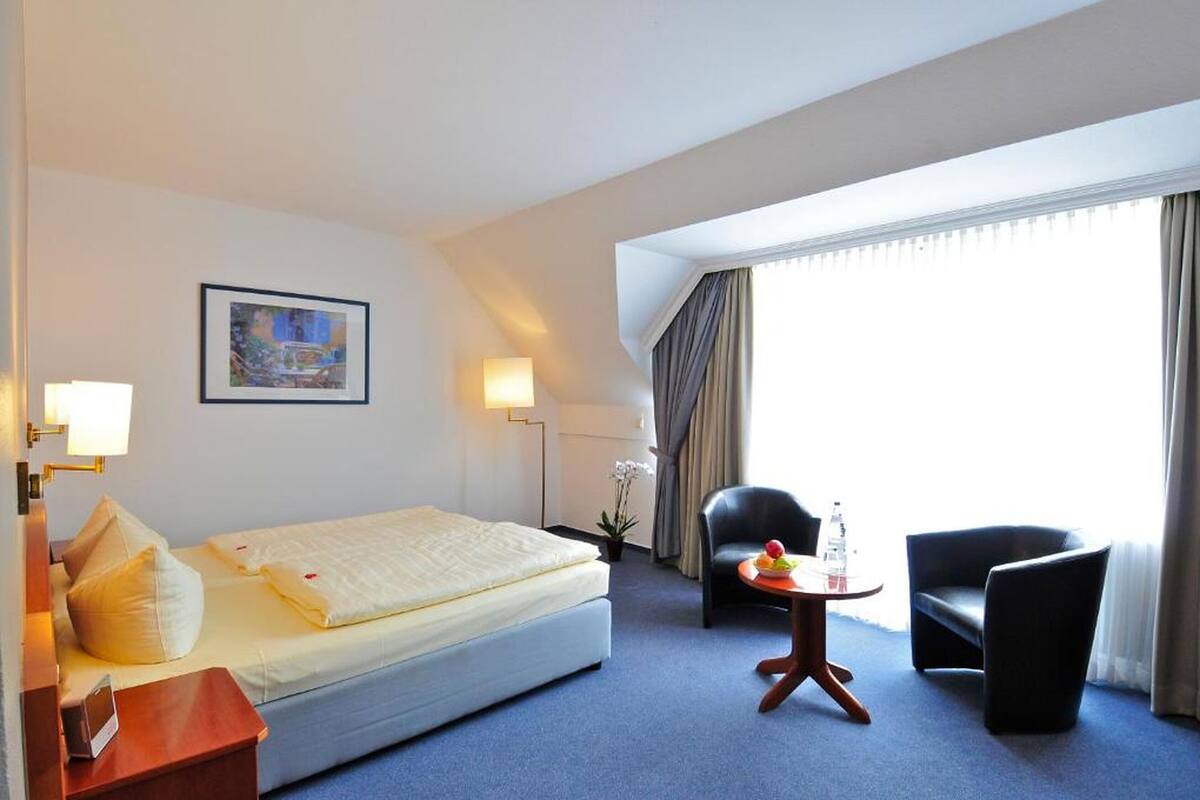 A hotel room with a bed, two chairs, a small table with fruit, a lamp, and a painting on the wall.