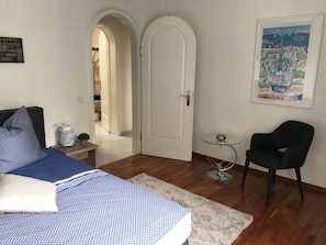 Un dormitorio con cama, una silla, una mesita y una puerta que da a otra habitación.