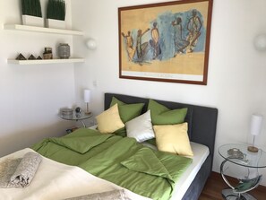 Una cama bien hecha con ropa de cama verde y blanca, una obra de arte enmarcada encima y una mesita de noche con una lámpara.