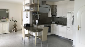 A modern kitchen with white cabinetry, a granite countertop, and a built-in oven. There are two chairs at the kitchen island, and a potted plant is visible on the left. - Villa Kaissini (Orselina)